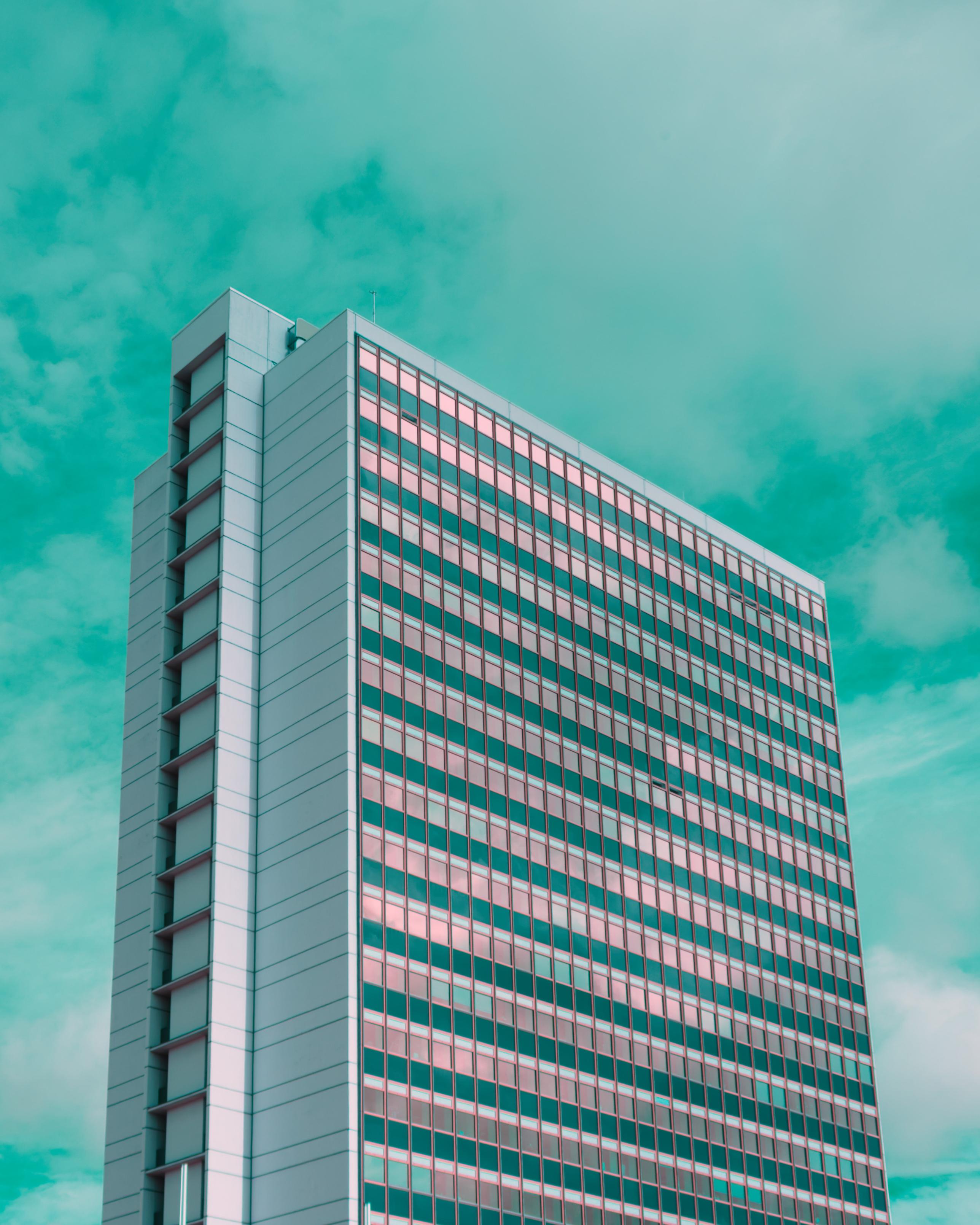 A photo up a weirdly iridescent office building. A few windows are open and reflect weirdly reddish. The sky is deeep vibrant turquoise.